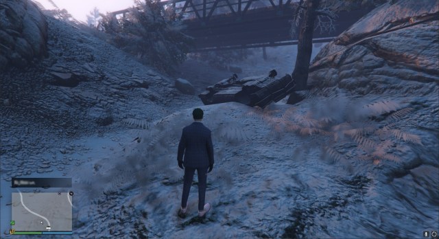 A GTA Online character with green hair and a navy suit standing beside a crashed car, on its roof, beside a tree. 