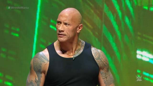 The Rock standing at the WrestleMania 40 press conference, wearing a necklace and a black tank top.